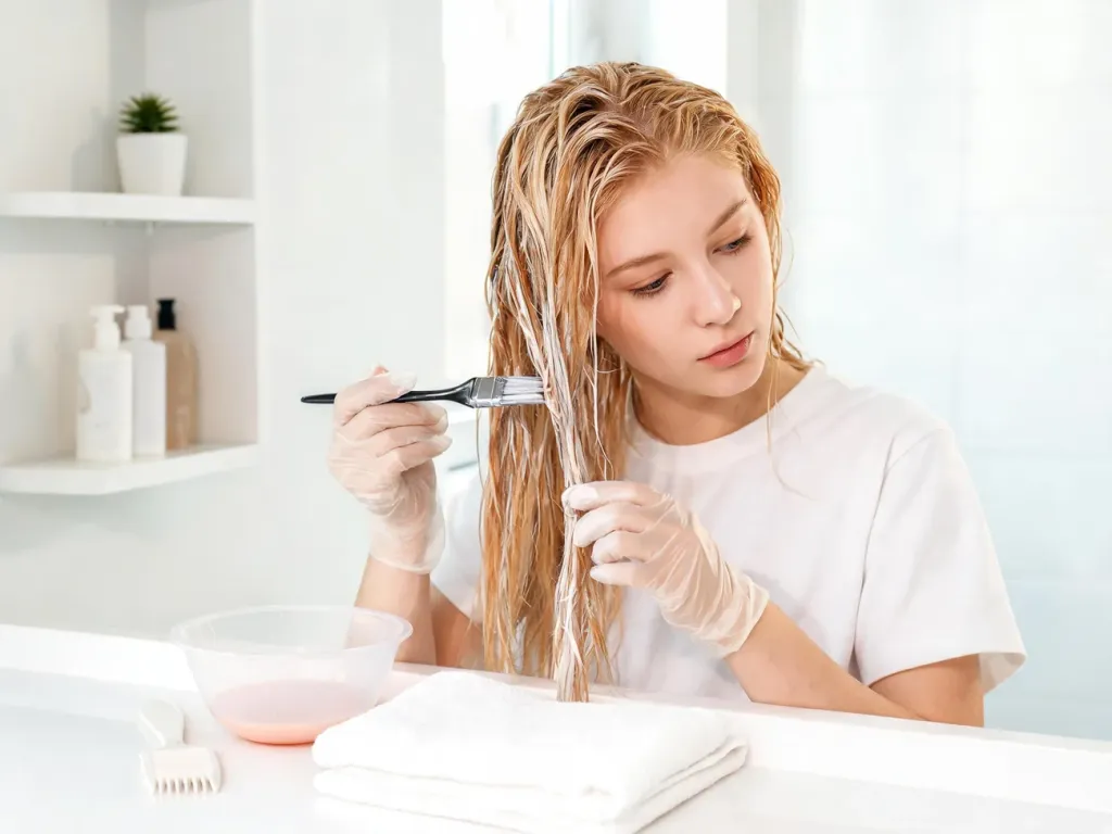 Młoda kobieta w jasnej łazience nakłada toner pędzelkiem na wilgotne blond włosy, obok miseczka z tonerem i grzebień.